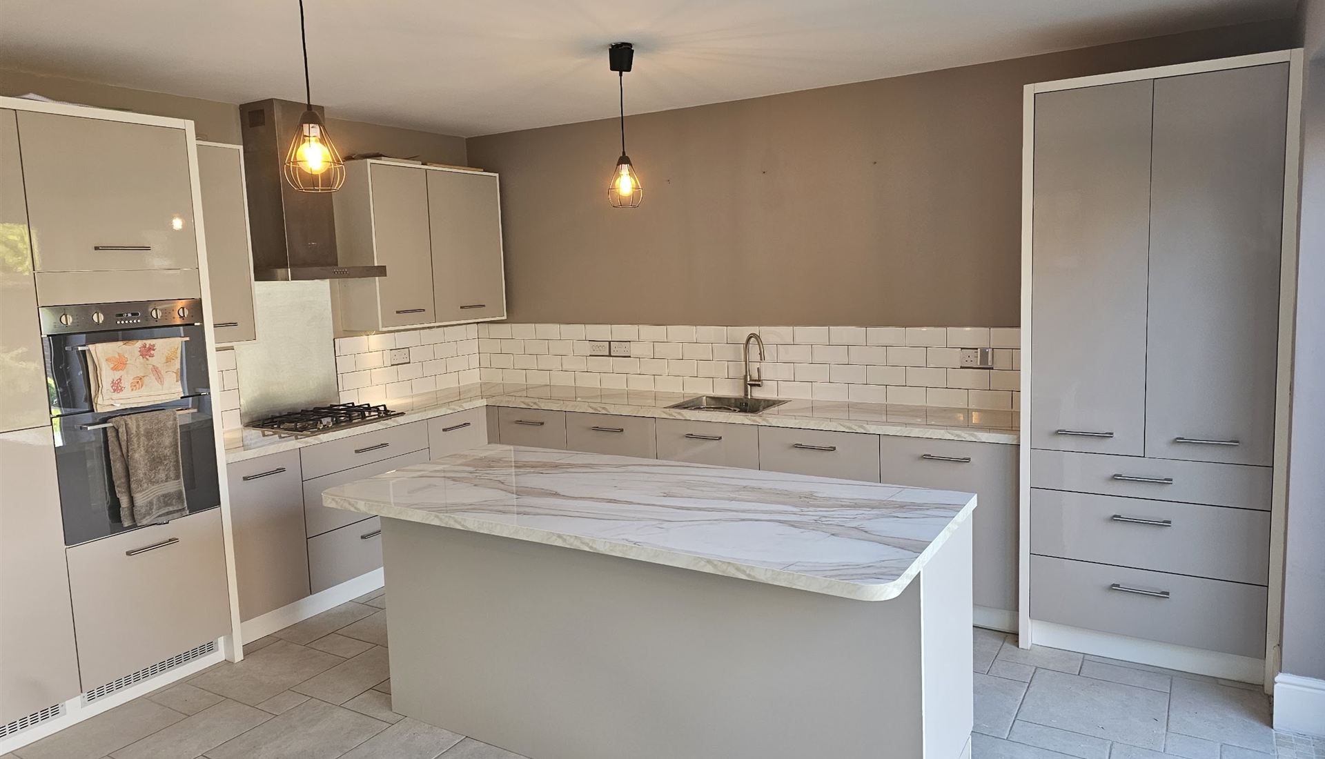 Finished kitchen with a marble-effect island, pendant lights and tall larder units, fitted by Amiry Renovation